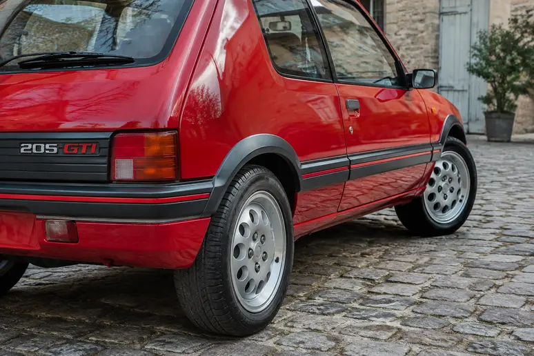 Peugeot 205 sportive rouge garée dans une cour, mettant en valeur son état d'origine.